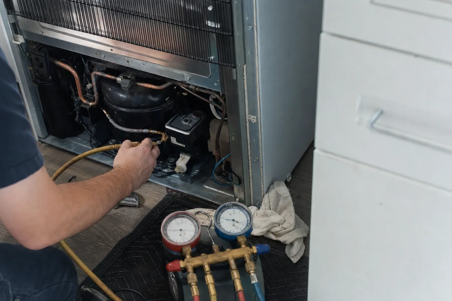 Refrigerator pulled out of its cabinet with the back panel off, a forearm holding a charging hose at the compressor and manifold gauges on the floor