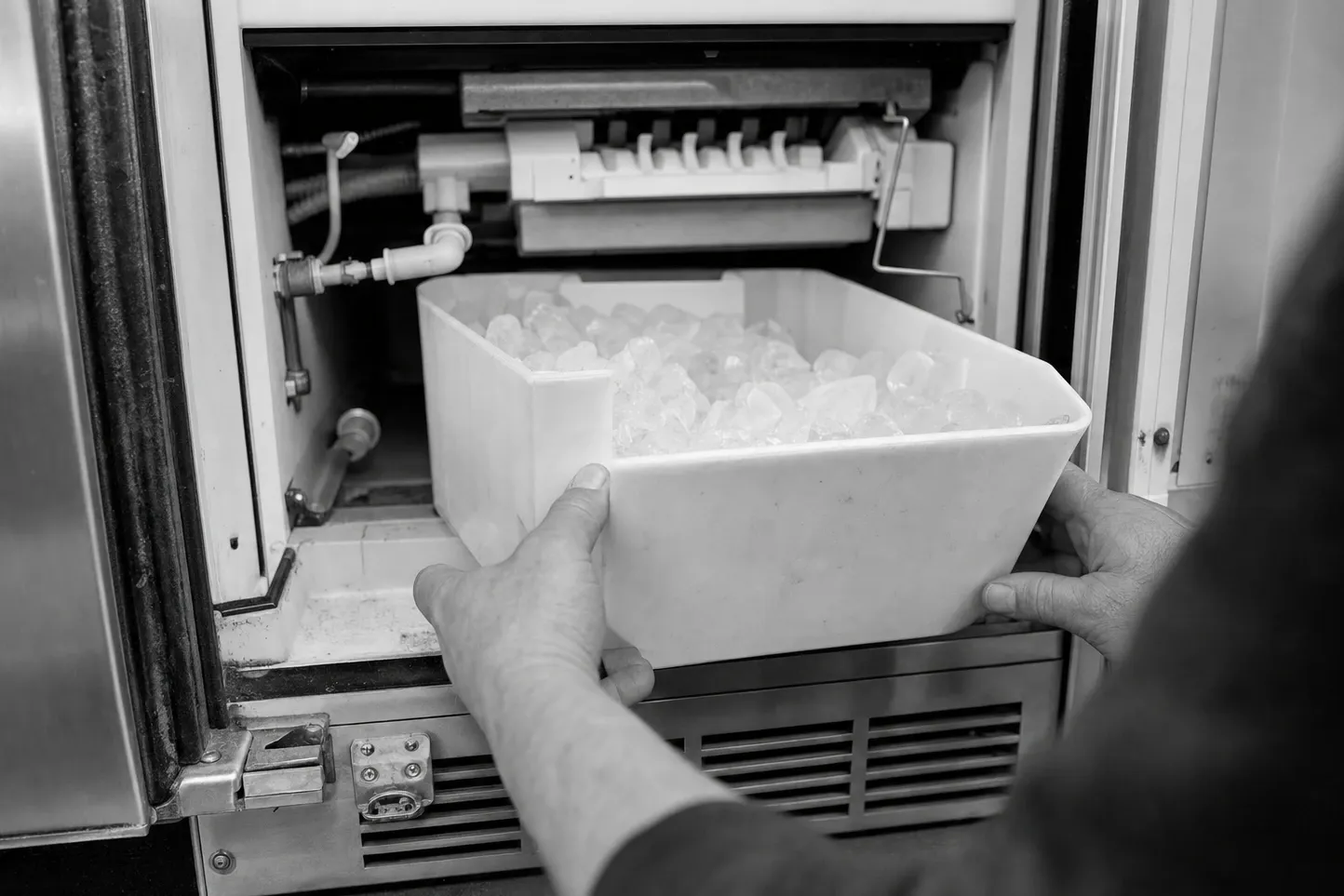 Black-and-white frame of two hands lifting a bin of ice cubes out of an undercounter ice maker, cube mould and fill plumbing above