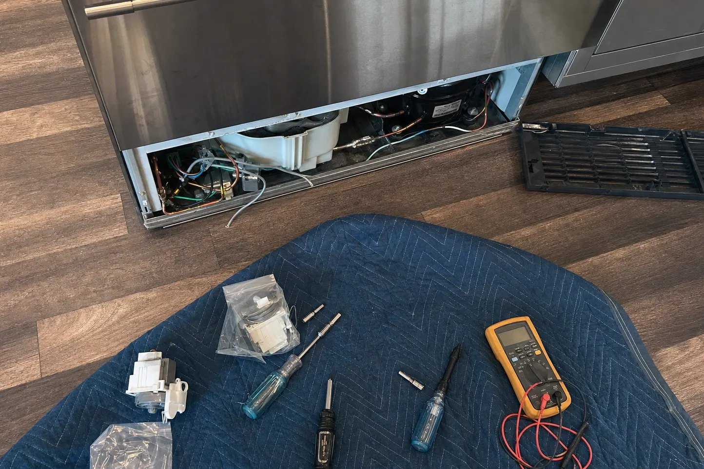 Lower grille removed from a tall stainless built-in and lying on the floor, exposing the compressor, white condenser-fan housing and copper lines, with a multimeter, drivers and bagged parts on a blanket