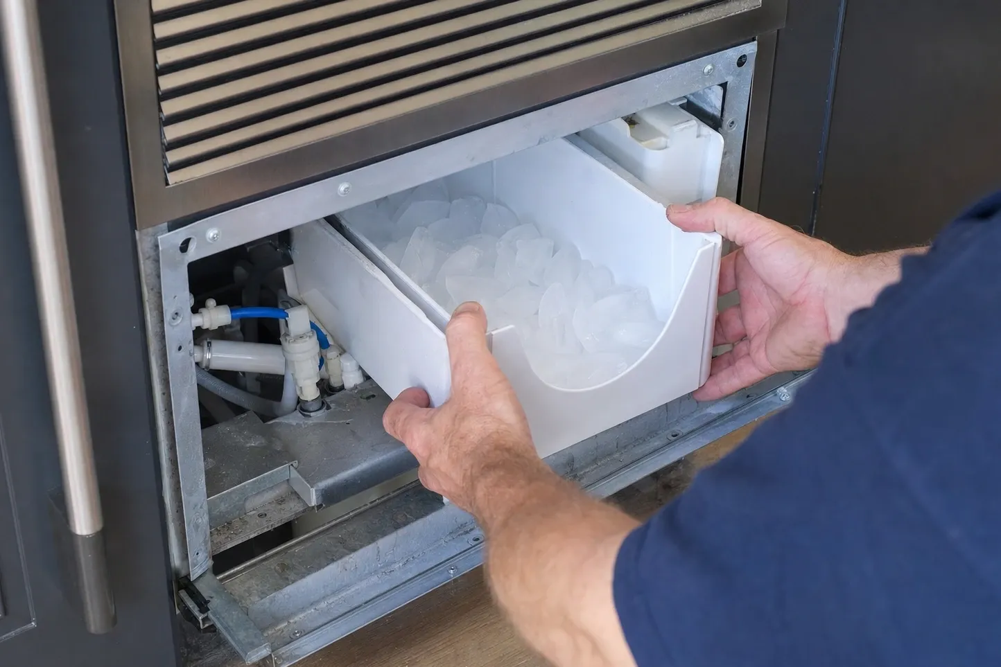 Hands drawing a white storage bin full of cubes out of a machine below a louvred grille, blue water tubing and white plumbing fittings visible in the bay to the left