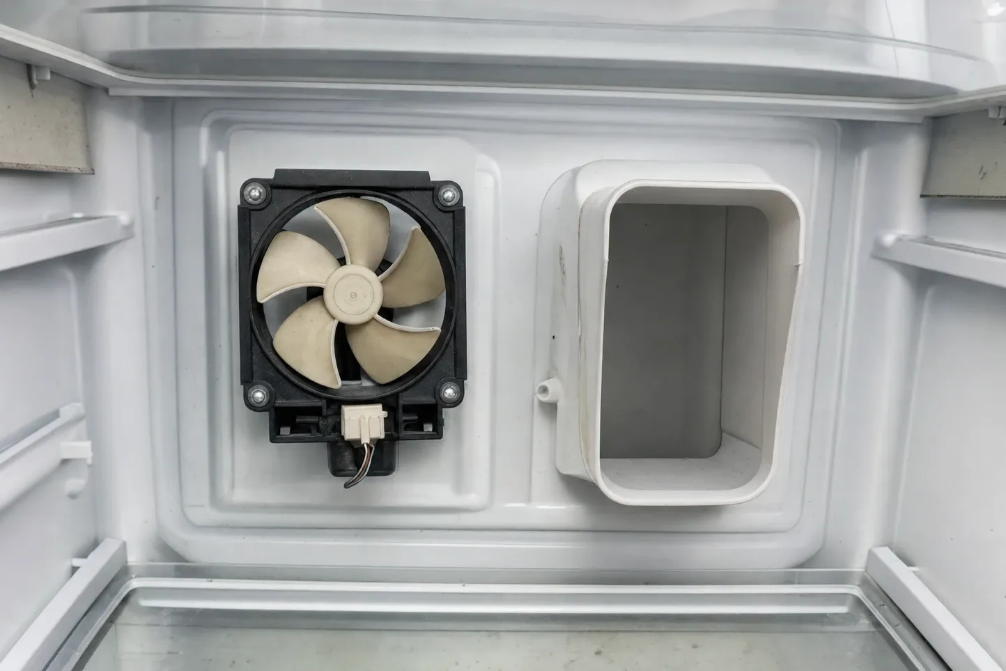 Five-bladed circulation fan in a black frame set into the rear wall of an empty white compartment, a moulded return-air duct beside it and a glass shelf underneath